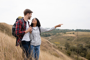 African american woman with backpack pointing with finger at landscape near boyfriend with backpack