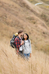 Smiling multiethnic couple with backpacks holding hands on grassy hill