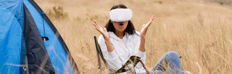 Excited african american woman using vr headset near tent and grass on blurred foreground, banner