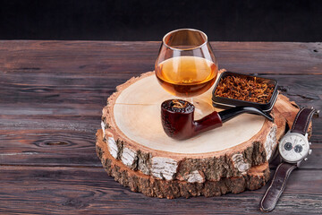 Obraz na płótnie Canvas Concept of time and unhealthy lifestyle. Glass of brandy and smoking pipe on a round wooden board.