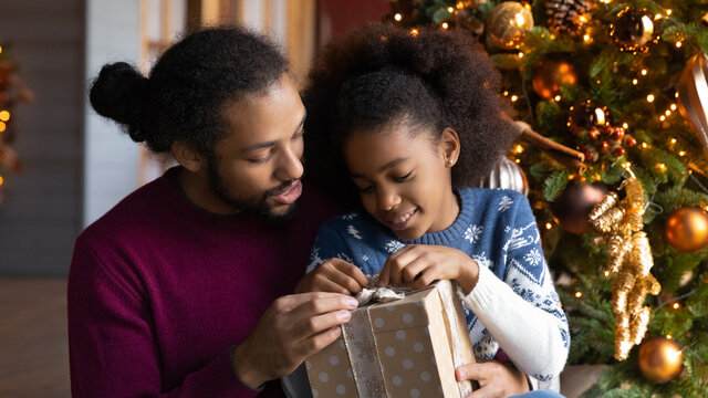Close up African American father and adorable daughter unpacking Christmas gift, sitting near festive tree at home, loving dad and little girl hugging, unwrapping present box, winter holiday