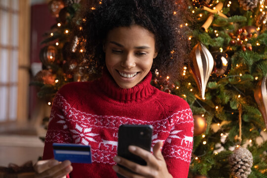 Close up smiling African American woman paying online by credit card for Christmas gifts, using smartphone, looking at screen, sitting near festive tree, shopping online, enjoying winter holiday