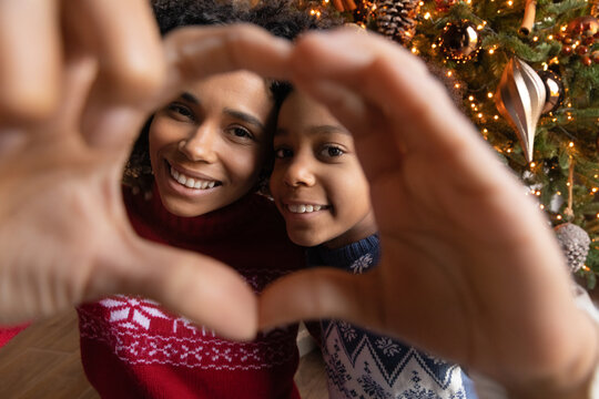 Close up African American mother and adorable daughter wearing sweaters showing heart sign, looking through fingers at camera, taking selfie near Christmas tree, celebrating New Year at home