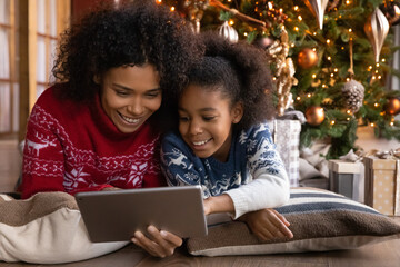 Close up happy African American woman with daughter using tablet together, lying on soft pillows near Christmas tree at home, happy family shopping online, choosing gifts, enjoying winter holiday - Powered by Adobe