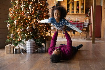 Portrait African American father lying on warm floor, carrying smiling adorable daughter, celebrating Christmas at home, loving dad and little girl playing active game, having fun on winter holiday