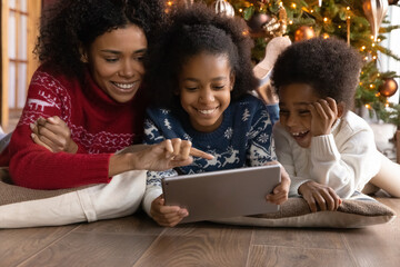 Close up happy African American family using tablet, lying on floor near Christmas tree, smiling mother with adorable daughter and son shopping or chatting online, enjoying winter holiday