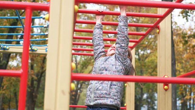 Little girl swinging on playground monkey bars hanging on arms in autumn park