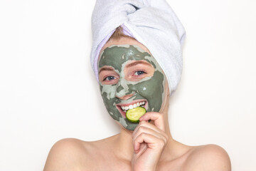 A young woman with a towel on her head and a clay mask on her face and a piece of cucumber. Skin care at home. White background.