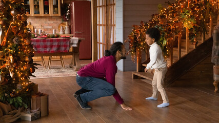 Obraz na płótnie Canvas Happy African American man playing funny active game with son, enjoying winter holidays at home, smiling father and adorable little boy dancing near festive Christmas tree, celebrating New Year
