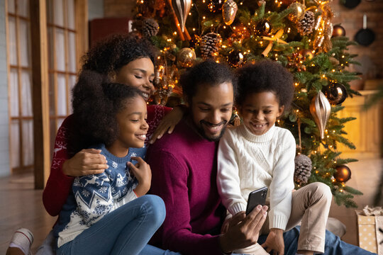 Happy African American family with two kids using smartphone, sitting near festive tree at home, smiling mother and father with son and daughter chatting or shopping online on Christmas holiday