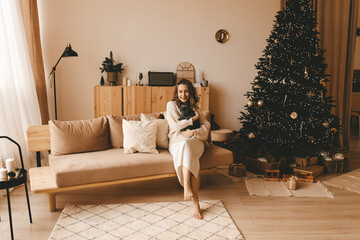 Happy girl in knitted cozy clothes hugs her cat sitting on the sofa near the decorated Christmas tree in the bright interior of the house. Concept of new year's shooting with your best friend