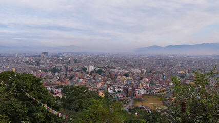 Panorama view of densely populated city center of Kathmandu, capital of Nepal, with visible smog viewed from Buddhist temple complex Swayambhunath with colorful prayer flags, trees and mountains.