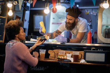 caucasian male employee in fast food service smiling and talking to a afro-american female customer while adding mustard in her sandwich