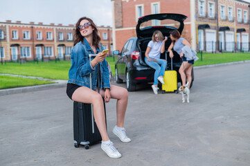 Fototapeta na wymiar Three girlfriends go on a road trip. Young woman posing with a suitcase. Two girls are sitting on the trunk of a car and watching a map.