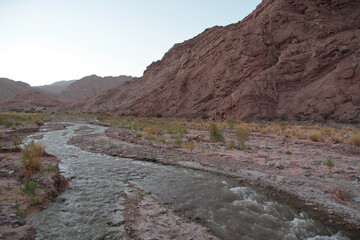 Magical landscape at sunset. Beautiful view of the pure water stream flowing across the desert, canyon and red sandstone mountains with golden hour colors. 