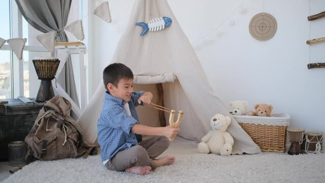 Little boy shooting with wooden slingshot sitting on floor in front of wigwam in children's room