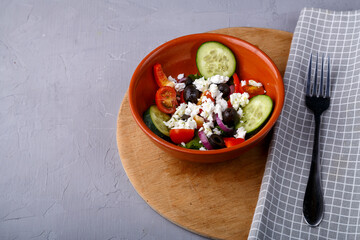 A brown plate of greek salad on a round stand next to a napkin and a fork on a gray concrete background. Copy space.