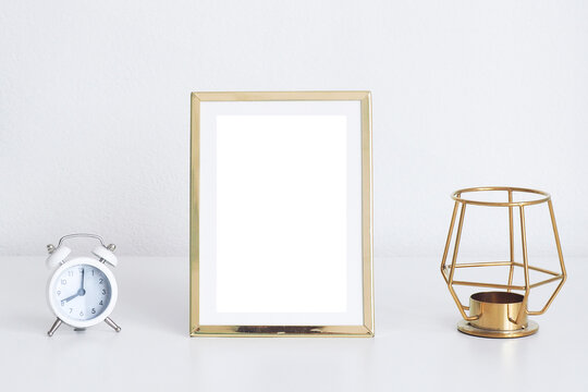 mockup gold photo frame, white alarm clock and gold candlestick on white table..