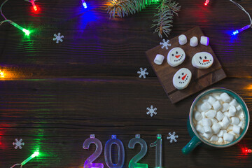 Christmas New Year card. Cup of hot drink with marshmallows, snowmen on a dark wooden background. Festive concept with snowflakes, colored garland and 2021 numbers. Top view. Copy space.