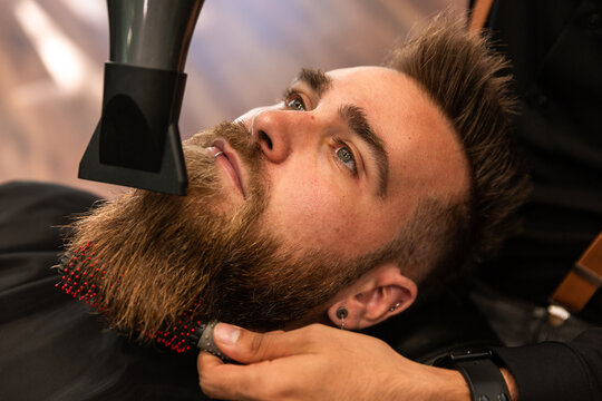 A bearded boy has his beard combed by a barber with a comb and a hairdryer.