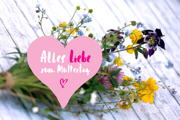 Fototapeta na wymiar Alles Liebe zum Muttertag . Weißer Hintergrund aus Holz mit pinken Herz, Karte mit Text, Handschrift und bunten, selbstgepflückten Blumen.