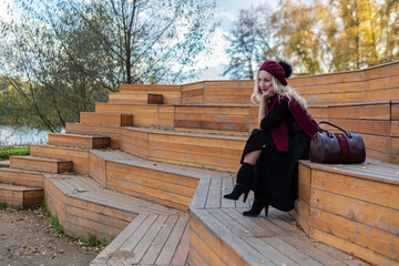 Fototapeta premium A happy girl sits on a wooden summer theater bench in a burgundy coat and biret, looking at the camera, in the fall against the backdrop of a pond with blue clouds.