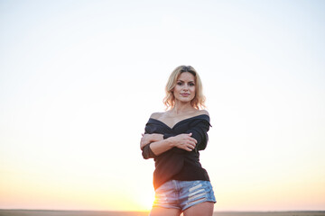 Obraz premium Young blond woman, wearing black jacket and jeans shorts, posing on wheat field on summer evening. Creative stylish three-quarter female portrait during sunset at natural rural landscape.