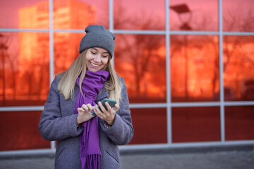 Happy casual excited woman celebrating success holding mobile phone standing on the street warmly dressed in a gray hat and a purple scarf near a shopping center on a bright red background