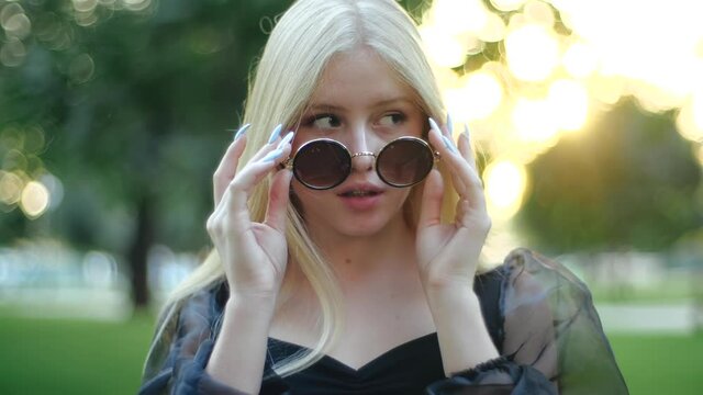 A caucasian girl takes off her round sunglasses and turns around in surprise. The young fashion model coquettishly and with interest looks at the camera and waves her long blond hair, posing.