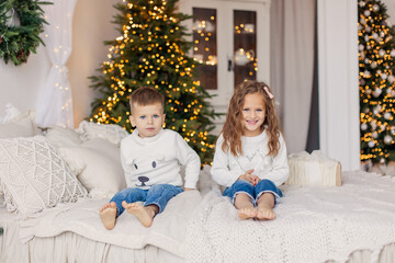 Obraz na płótnie Canvas Siblings brother and sister are lying in bed. Christmas morning cozy bedroom. Light Scandinavian interior. Children are cheerful happy in light sweaters. Childhood game fun. New Year European 