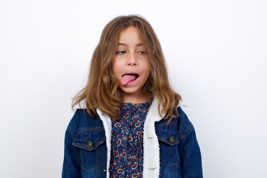 Little caucasian girl with beautiful blue eyes wearing denim jacket standing over isolated white background showing grimace face crossing eyes and showing tongue. Being funny and crazy