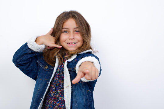 Little Caucasian Girl With Beautiful Blue Eyes Wearing Denim Jacket Standing Over Isolated White Wall Smiling Cheerfully And Pointing To Camera While Making A Call You Later Gesture, Talking On Phone