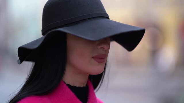 Close-up of a stylish black-haired woman in a pink coat and black hat walking on a blurred background of a city street.