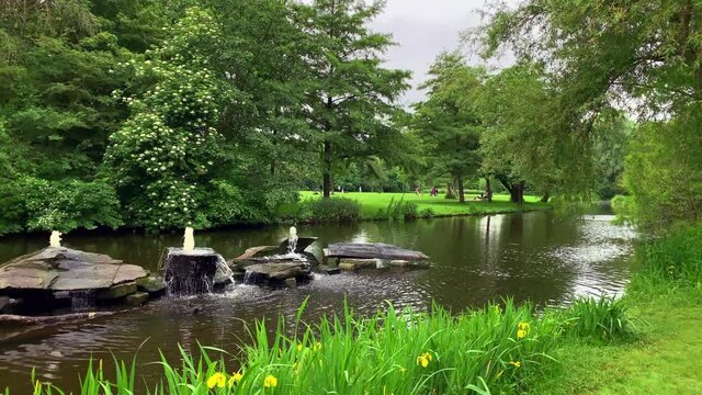 Footage of people walking, small water fountains from rocks, lake, trees at Vondelpark in Amsterdam. It is a sunny summer park day. Camera moves forward.