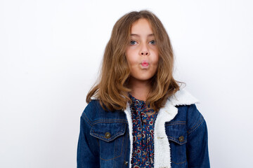 Shot of pleasant looking Little caucasian girl with beautiful blue eyes wearing denim jacket standing over isolated white background pouts lips, looks at camera, Human facial expressions