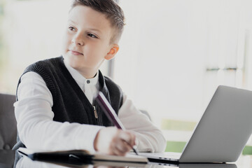 makes lessons online with a teacher and writes them in a notebook. dressed in a school white shirt and a stylish hairstyle studying on a laptop looking at the screen
