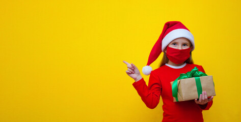 A girl in a Santa hat, in a red T-shirt, wearing a medical mask in her hands holds a gift for Christmas and New Year and points her finger towards the place for advertising. 