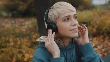 Young blond woman listening to the music from her smartphone in the park using the headphones. Portrait. High quality 4k footage