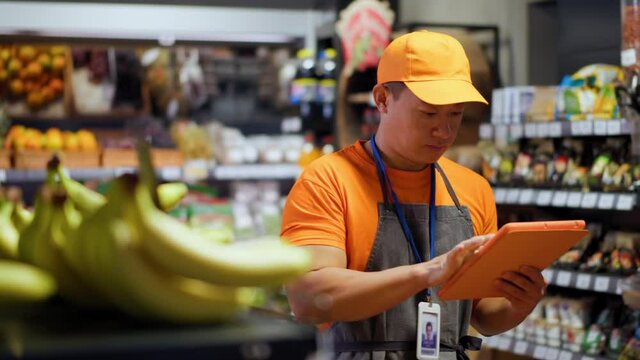 Asian shopkeeper employee using digital tablet distributing arranging fresh fruits vegetables on shelves. Professional workman. Chinese ethnicity. Supermarket foods. Retail store.