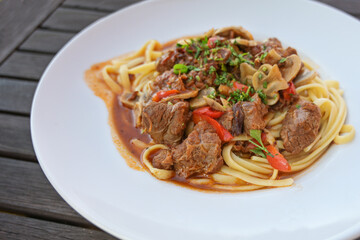 Ragout from beef, red bell peppers and mushrooms with parsley garnish on pasta in a white plate on a dark rustic wooden table, copy space, selected focus