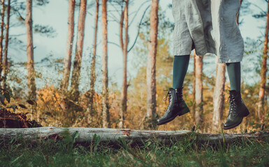 Side view of woman dressed in grey dress, green tights and black boots jumping from a tree log with pine tree forest in autumn background