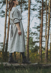 Full body side view portrait of caucasian young woman in a long organic linen dress, with pine tree forest in the background. Concept: Slow fashion, minimalism, handmade clothes, natural beauty