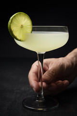 Man's hand grabbing a classic Daiquiri cocktail with a lime wheel from a black table
