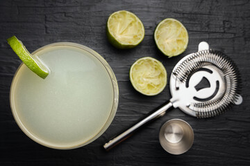 Top view of a classic Daiquiri cocktail with a lime wheel. Squeezed limes, strainer and jigger over a black table