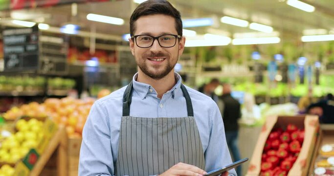 Cheerful smiling Caucasian handsome male in glasses standing at market and typing on tablet. Young joyful man in apron at work browsing on device indoors. Close up portrait. Business concept