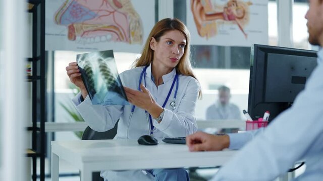 Doctor woman sitting at table in medical office looking at x-ray scan of lunges and discussing diagnosis with man patient during consultation. Pneumonia diagnose. Slow motion