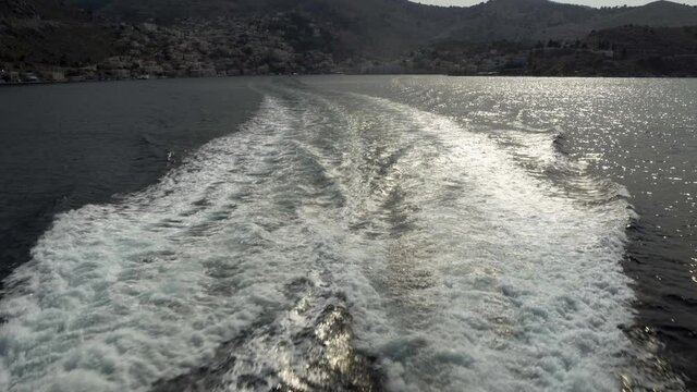 Chalki / halki island, Greece: Waves formed by ship sailing away from island situated on a rusty hills during day time. Currents formed on a surface of water due to motion of ship