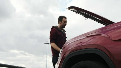 man in a red overalls on the street arrived on call from a client, he opens the hood of burgundy SUV and tries to fix the breakdown. Looks under the hood.