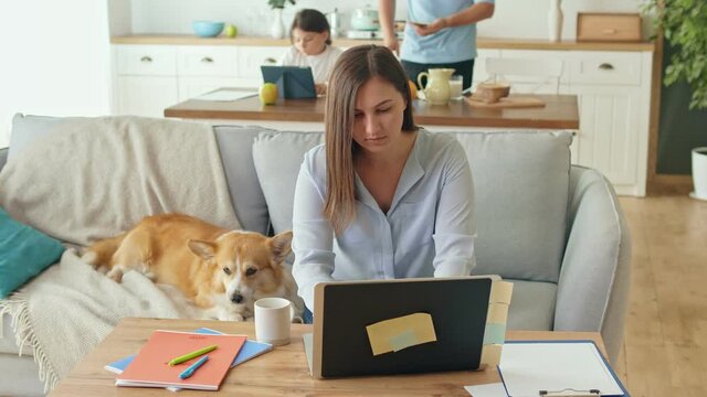 Busy Mom Working Remotely with Laptop Sitting on the Couch. Working From Home During a Quarantine Epidemic. In the Background, Her Daughter is Using a Tablet, her Husband is Busy in the Kitchen