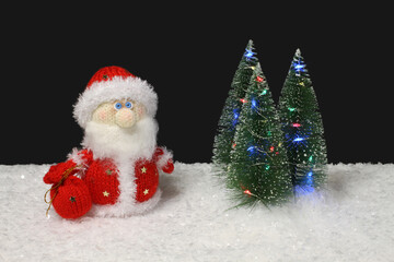 The toy knitted wool Santa Claus stands next to the toy green Christmas trees with lights on artificial white snow against black background. Closeup